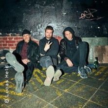 Drei Personen sitzen entspannt auf dem Boden vor einer dunklen Wand mit Graffiti.