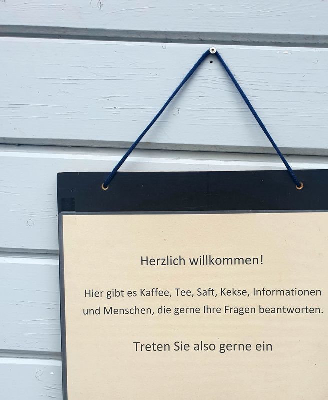 Ein Schild an einer Wand mit der Aufschrift: 'Herzlich willkommen! Hier gibt es Kaffee, Tee, Saft, Kekse, Informationen und Menschen, die gerne Ihre Fragen beantworten. Treten Sie also gerne ein.'