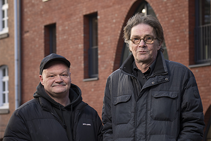 Foto von den Hinz & Kunzt Guides Chris und Uwe. Beide stehen nebeneinander vor einem Backsteinhaus, tragen warme Jacken und schauen mit einem freundlichen Blick in die Kamera..