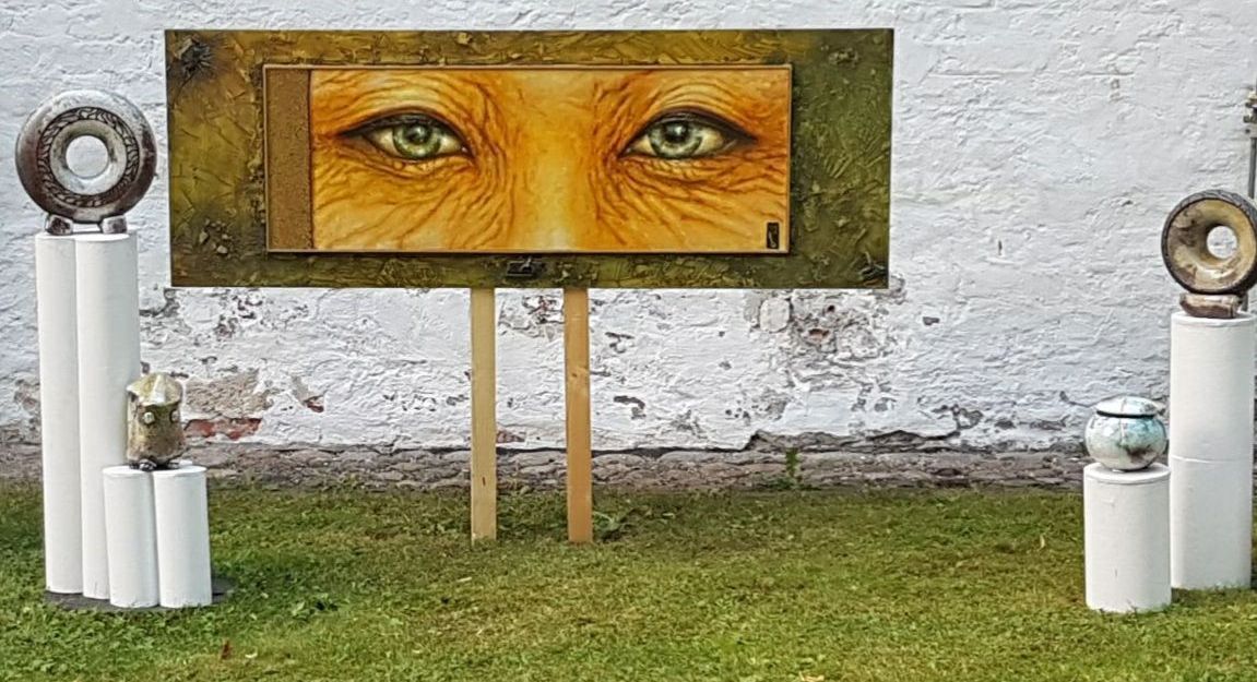Kunstinstallation mit einem Gemälde von Augen und Keramikobjekten auf weißen Säulen im Freien.