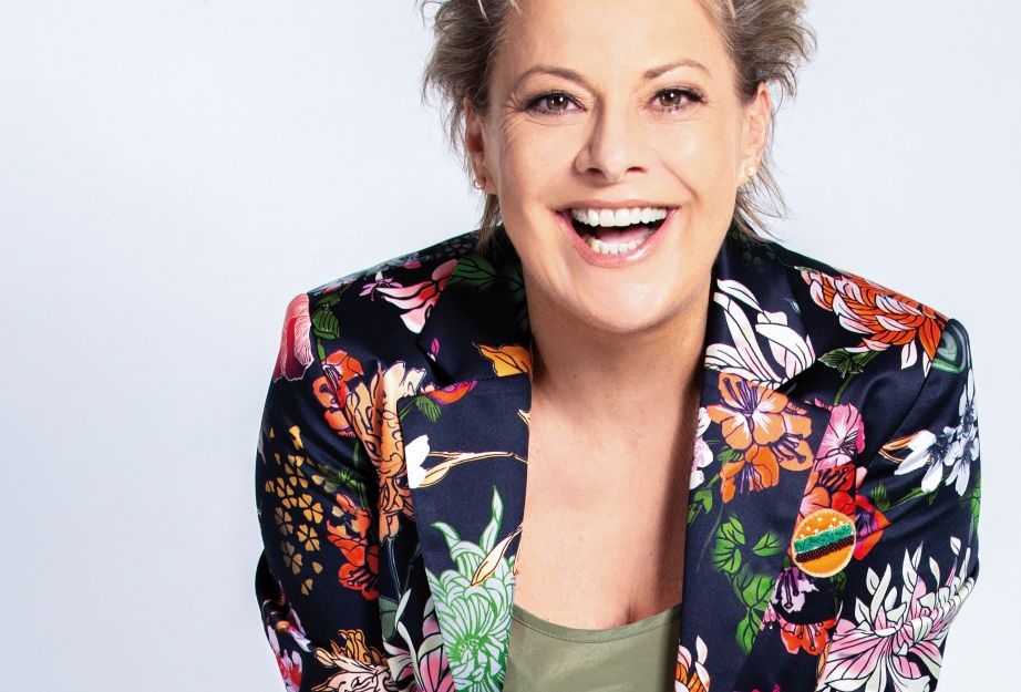 Eine lächelnde Person in einem bunten, geblümten Blazer vor einem hellen Hintergrund.