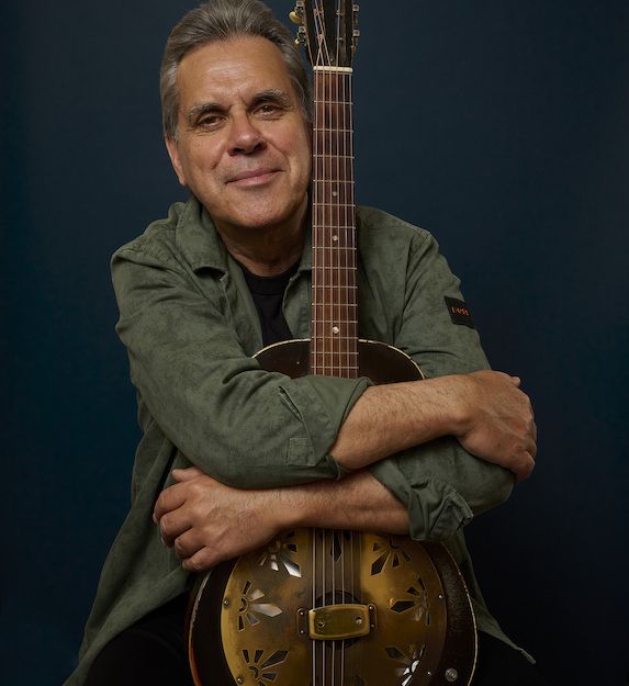 Ein Mann umarmt eine Gitarre vor einem dunklen Hintergrund.