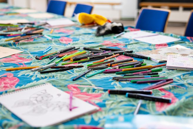 Ein Tisch mit vielen bunten Stiften und Malbüchern auf einer tropisch gemusterten Tischdecke.