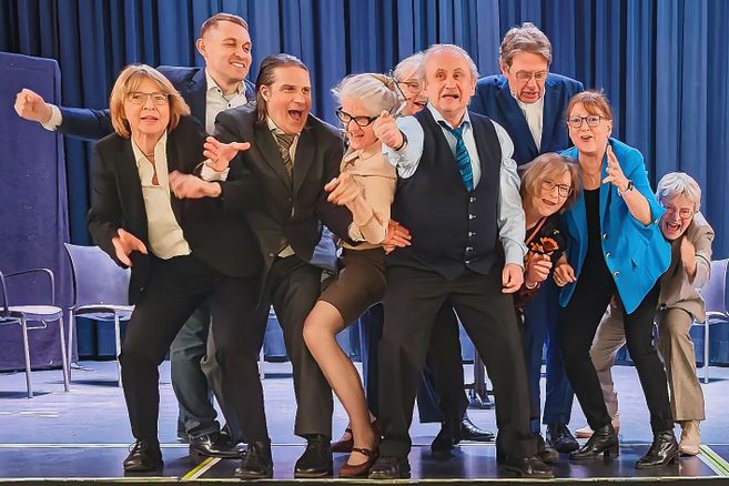 Ensemble von acht Personen auf einer Theaterbühne vor blauem Vorhang, in dynamischer Pose und offenbar mitten in einer Aufführung.