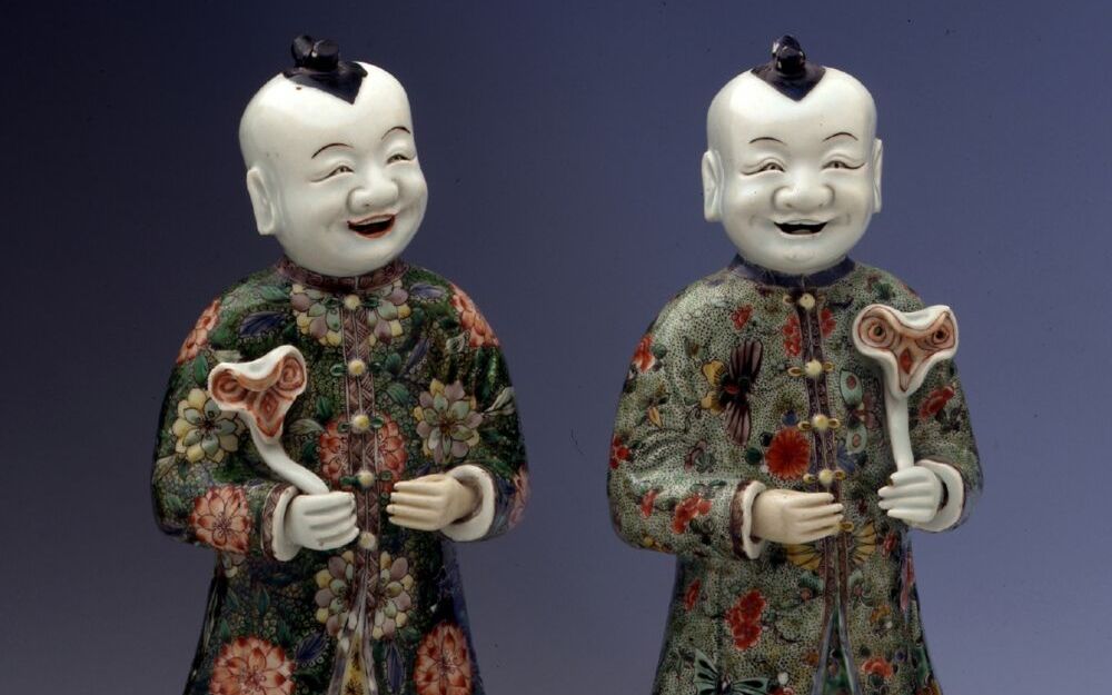 Zwei freundlich lächelnde Porzellanfiguren. Sie wirken männlich, ihr Gewand besteht aus Rock und Mantel, verziert mit chinesischen Mustern. Beide haben glattrasierte Köpfe mit einem Haarstreifen in der Mitte und halten etwas in der Hand.