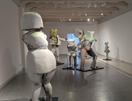 A piece of art in an exhibition space with several sculptural figures. The figures have different colors and sizes and stand on black pedestals.