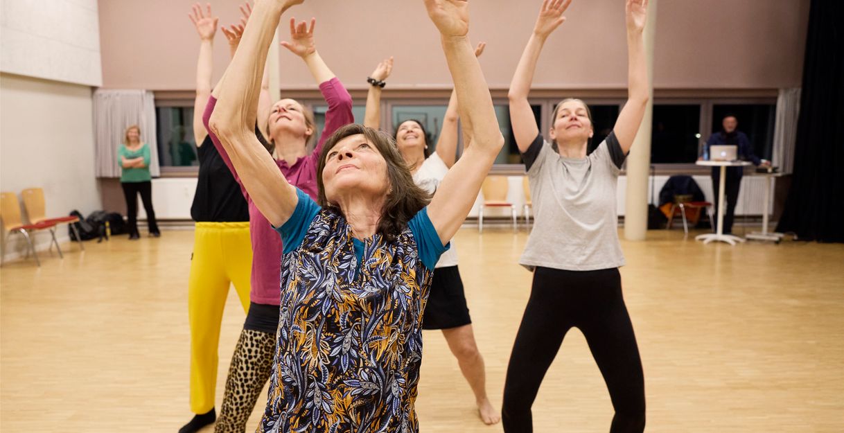 Five people are standing behind each other in a dance studio, each with both arms stretched toward the ceiling. They are wearing colorful sportswear and their eyes are directed toward the ceiling.