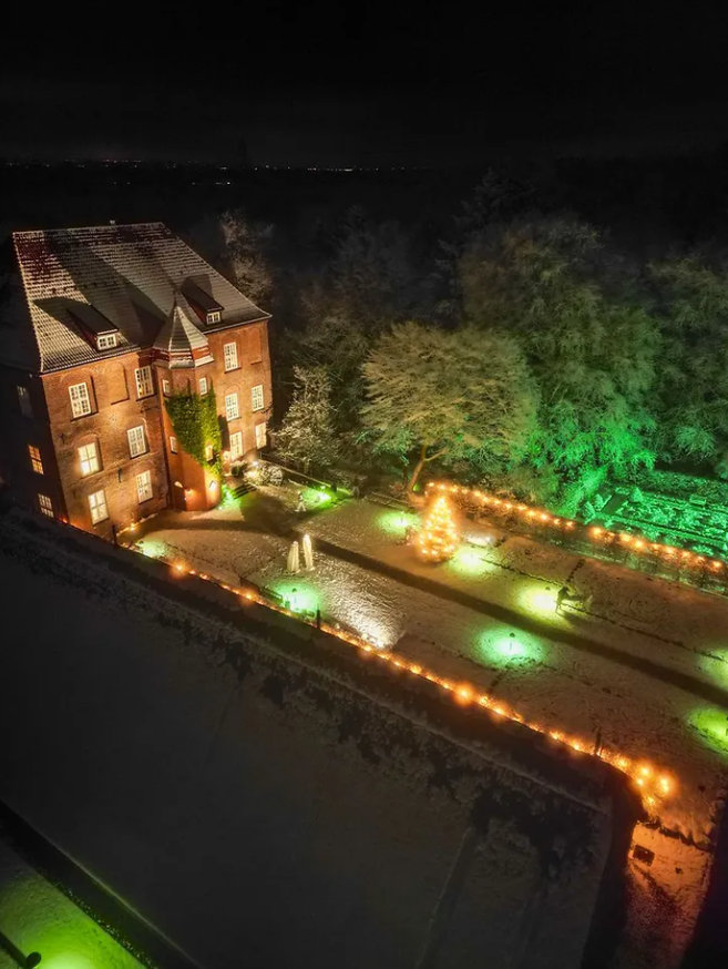 Nachtaufnahme eines beleuchteten Hofs am Weihnachtsmarkt für Kunsthandwerk & Design mit Backsteingebäude, Lichterketten und geschmücktem Baum im Schnee.