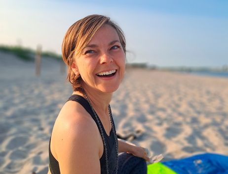 Frau sitzt lächelnd auf einer bunten Decke am Strand.