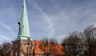 Kirche St. Johannis in Hamburg-Eppendorf mit spitzem Turm und rotem Dach, umgeben von Bäumen.