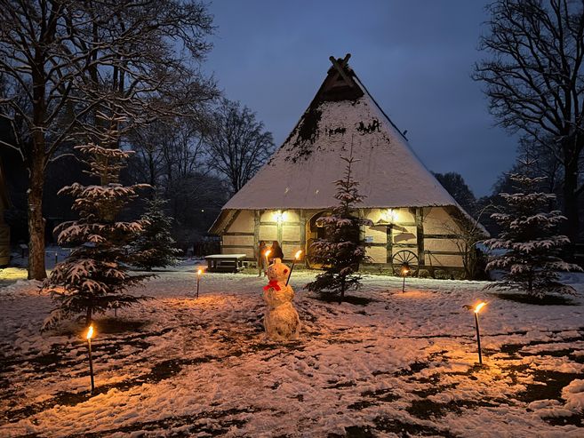 Ein verschneites Fachwerkhaus mit beleuchtetem Eingang, umgeben von Tannenbäumen und Fackeln. Ein Schneemann steht im Vordergrund.