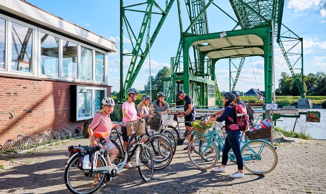 Gruppe von Radfahrern mit Helmen steht vor einer grünen Brücke und einem Backsteingebäude.