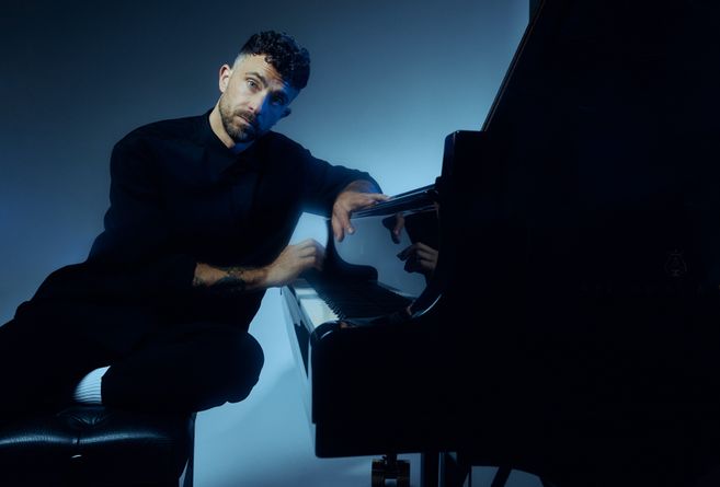 Ein Mann in dunkler Kleidung sitzt an einem Flügel in einem stimmungsvoll beleuchteten Raum.