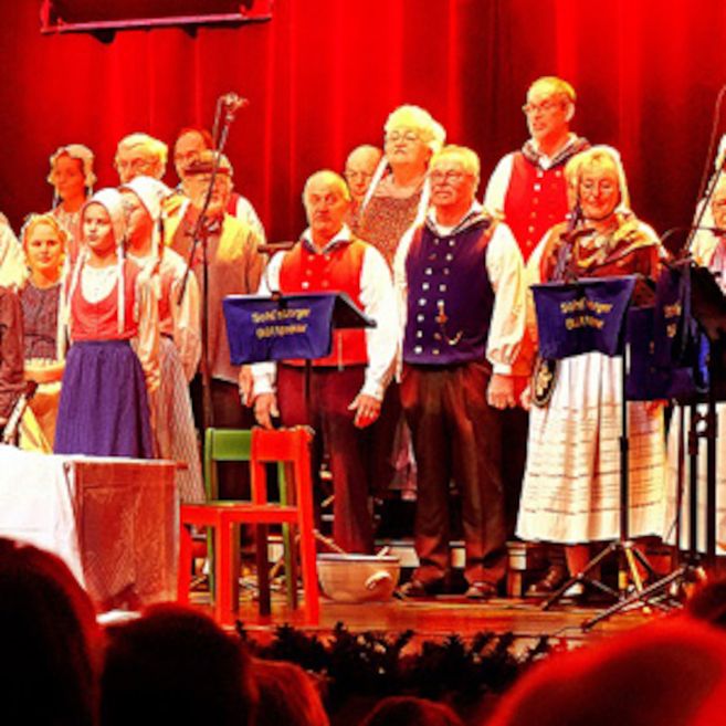Eine Gruppe von Menschen in traditioneller Kleidung singt auf einer Bühne vor einem roten Vorhang.