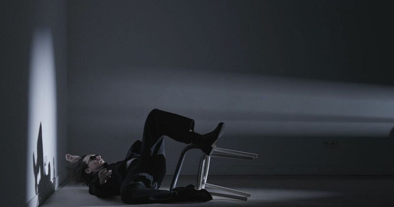 Person in dunkler Kleidung liegt auf dem Boden neben einem umgekippten Stuhl in einem dunklen Raum.