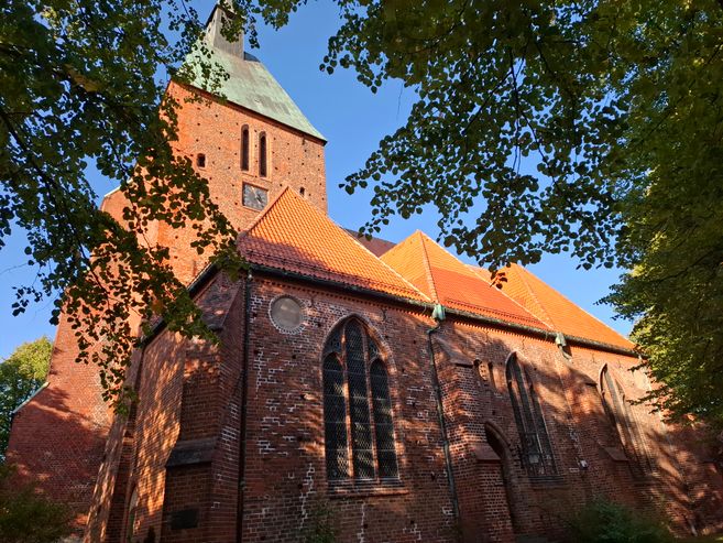 Backsteinkirche mit rotem Dach und Turm, umgeben von Bäumen.