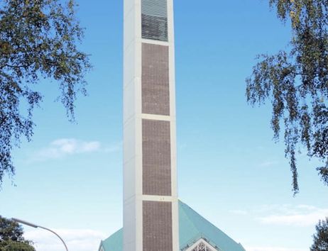 Moderne Kirche mit hohem Turm und grünem Dach, umgeben von Bäumen und einem blauen Himmel.