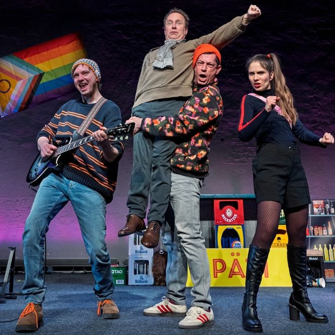 Vier Personen auf einer Bühne, eine Person spielt Gitarre, eine wird hochgehoben, eine trägt bunte Kleidung und eine posiert energisch. Im Hintergrund eine Regenbogenflagge.