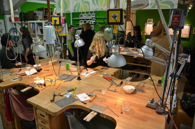ein großer Goldschmiedewerktisch auf dem sich Werkzeuge und Gläser befinden und im Hintergrund stehende Kursteilnehmende