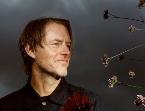 Ein Mann mit mittellangem Haar und Bart schaut lächelnd zur Seite. Im Vordergrund sind Blumen zu sehen, der Hintergrund ist dunkel und wolkig.