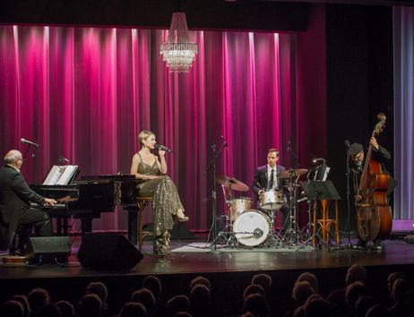 Eine Sängerin und ihre Band treten auf einer Bühne mit pinkem Vorhang auf. Ein Pianist, ein Schlagzeuger und ein Kontrabassist begleiten sie.