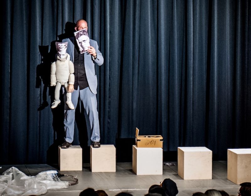 Ein Performer steht auf einer Bühne mit einer Puppe und hält ein Bild hoch, umgeben von schwarzen Vorhängen und Holzkisten.