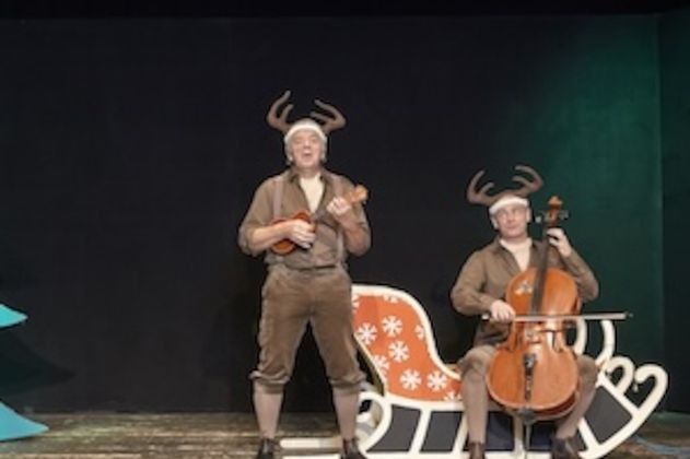 Zwei Männer in Rentierkostümen auf einer Bühne, einer spielt Ukulele, der andere Cello, neben einem Schlitten.