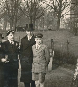 Schwarz-weiß Foto von vier Personen, die in einer parkänlichen Umgebung stehen. Von links: Frau mit feinem Mantel, junger Mann in Anzug, Mann mit Mantel und Zylinder, Junge mit Jackett und kurzen Hosen.