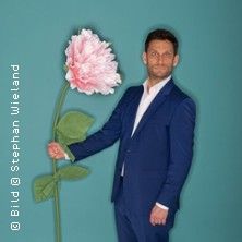 Ein Mann im Anzug hält eine übergroße rosa Blume vor einem blauen Hintergrund.