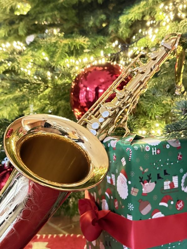 Ein Saxophon liegt vor einem geschmückten Weihnachtsbaum mit Lichtern und roten Kugeln. Daneben steht ein Geschenk mit Weihnachtsmotivpapier.