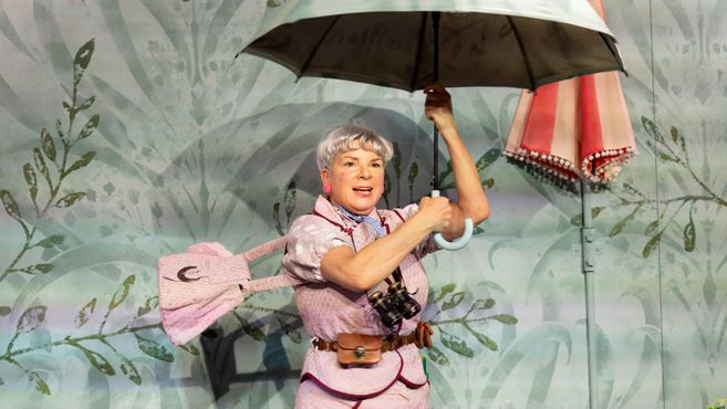 Frau mit kurzem grauem Haar hält einen geöffneten Regenschirm und trägt ein rosa Kleid mit passender Tasche vor einer Wand mit Pflanzenmuster