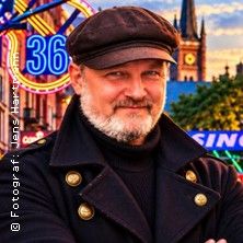 Porträt eines bärtigen Mannes mit Schirmmütze und dunklem Mantel vor der Reeperbahn in Hamburg; im Hintergrund leuchten das Schild „36“ und ein Kirchturm.
