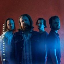 Vier Männer stehen vor einem roten Hintergrund, teils in blauem Licht beleuchtet.