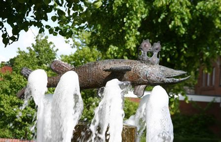 Bronzeskulptur eines Fisches mit Krone auf einem Brunnen, umgeben von sprudelndem Wasser und grünen Bäumen im Hintergrund.