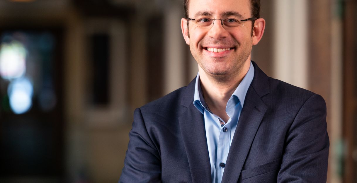 Ein Mann in einem blauen Anzug lächelt in die Kamera, während er die Arme verschränkt. Der Hintergrund ist unscharf und zeigt einen Innenbereich.