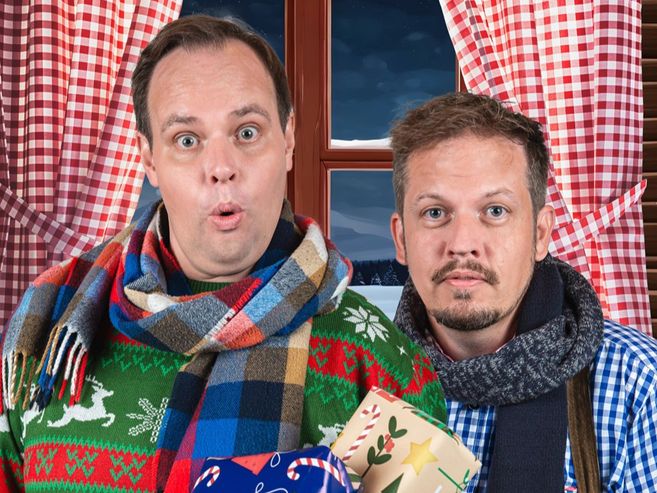 Zwei Männer in Weihnachtskleidung stehen vor einem Fenster mit rot-weiß karierten Vorhängen und halten Geschenke. Der Titel lautet „Komödie: Eine schöne Bescherung“.