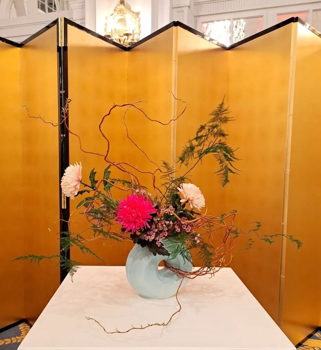 Blumenarrangement für den Geburtstag des Kaisers 2026, vor einem goldenen Wandschirm. Das Gesteck besteht aus 3 pinken und rosa Chrysanthemen, Korkenzieherhasel-Äste und weiteren Pflanzen.