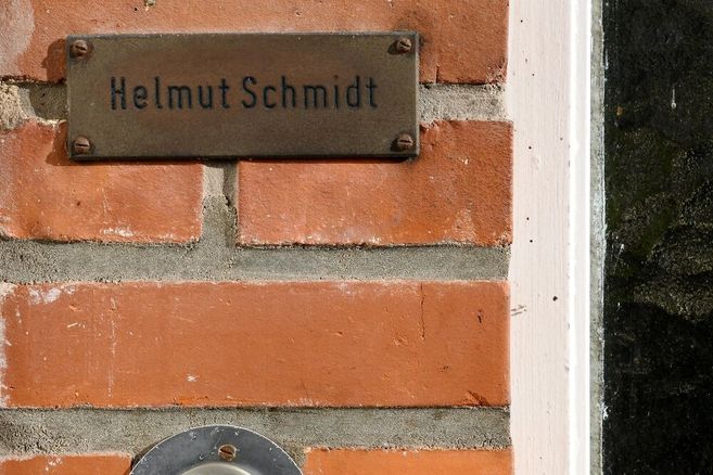Foto eines kleinen Ausschnittes einer Hauswand aus Backstein, in die eine runde Klingel mit Klingeknopf eingelassen sowie ein Metalschild befestigt ist. Auf dem Schild steht "Helmut Schmidt".