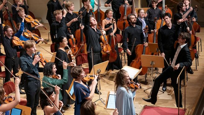 Ein Orchester auf einer Bühne, Musiker halten ihre Instrumente hoch und lächeln.