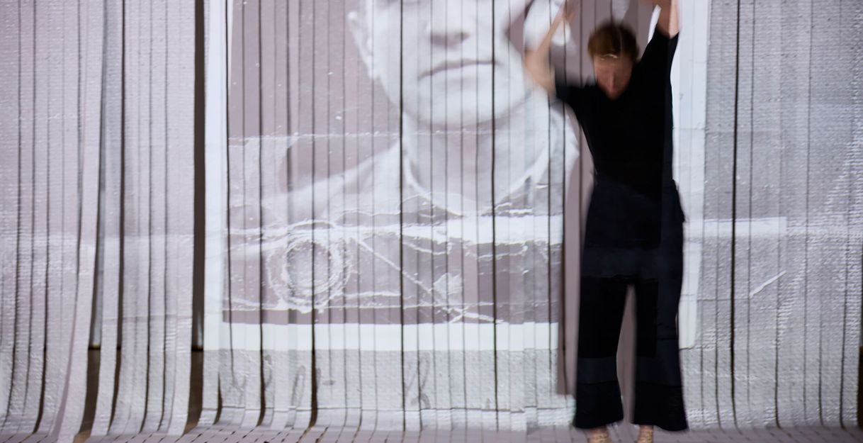 Die performende Person bewegt sich mit erhobenen Armen im Raum. Dahinter ist ein Vorhang aus weißen Streifen, auf den das Führerscheinbild eines Mannes zu sehen ist.