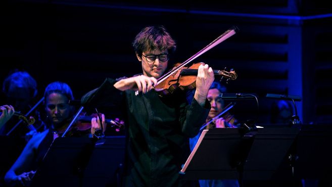 Ein Geiger spielt konzentriert auf der Bühne, umgeben von anderen Musikern im Hintergrund.