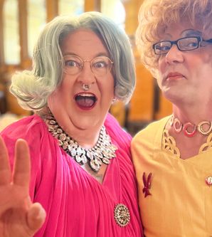 Zwei Dragqueens in bunten Outfits posieren fröhlich in einem eleganten Raum.