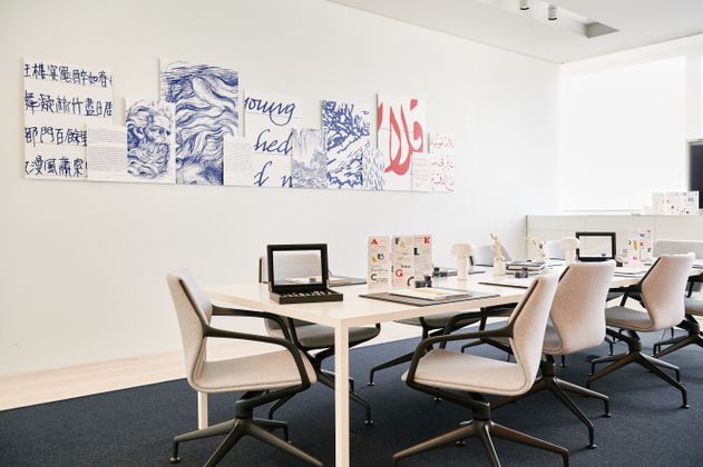 Tisch in der Mitte mit Schreibutensilien drauf, mehrere Stühle drum herum und Bilder an der Wand.