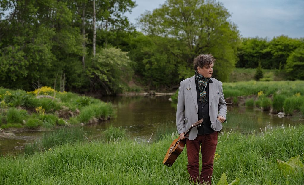 Ron Sexsmith stands in a green landscape holding his guitar.