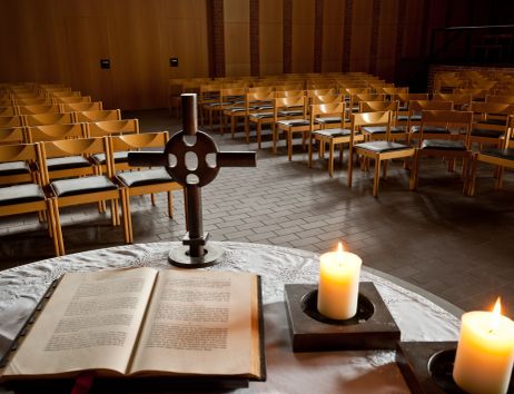 Ein leerer Kirchenraum mit einem Altar im Vordergrund, auf dem eine Bibel, ein Kreuz und zwei brennende Kerzen stehen.