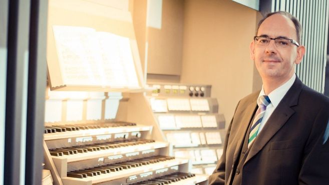 Ein Mann im Anzug steht neben einer Orgel mit mehreren Tastenreihen und Notenblättern.