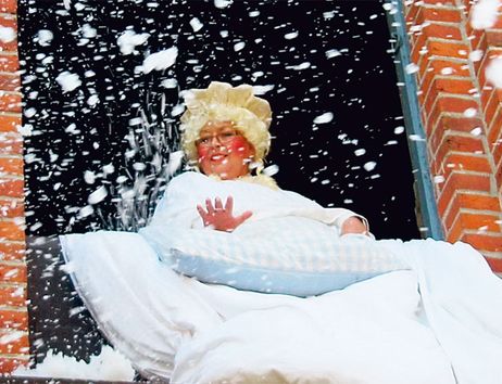 Eine Person in einem Frau Holle Kostüm schüttelt Kissen aus einem Fenster eines Backsteingebäudes, wodurch es schneit.