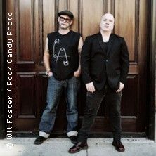 Zwei Männer stehen vor einer Holztür, einer trägt ein ärmelloses Shirt und Mütze, der andere einen Anzug.