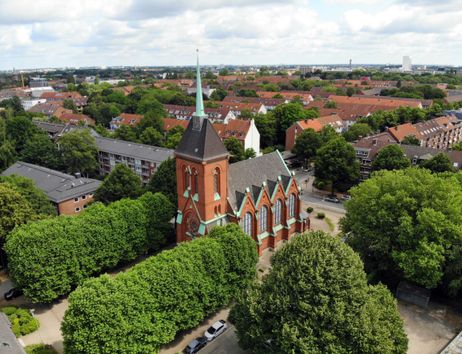 Luftaufnahme einer Kirche mit rotem Backstein und grünem Dach, umgeben von Bäumen und Wohnhäusern.