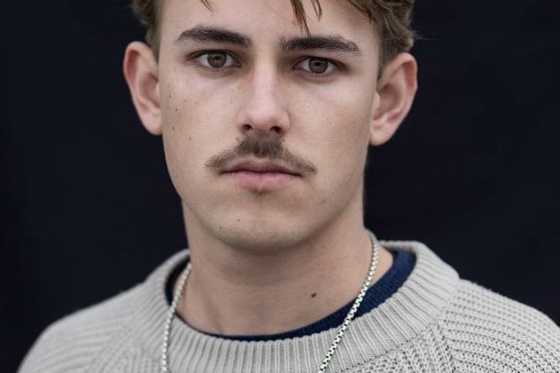 Porträt eines jungen Mannes mit Schnurrbart und Kette vor schwarzem Hintergrund.